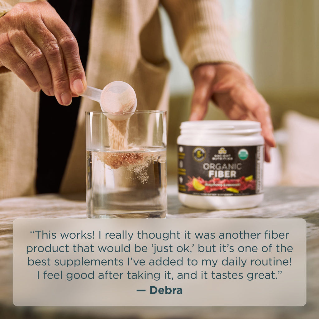 Person adding fiber powder to a glass with a container labeled 'Organic Fiber' on a table.