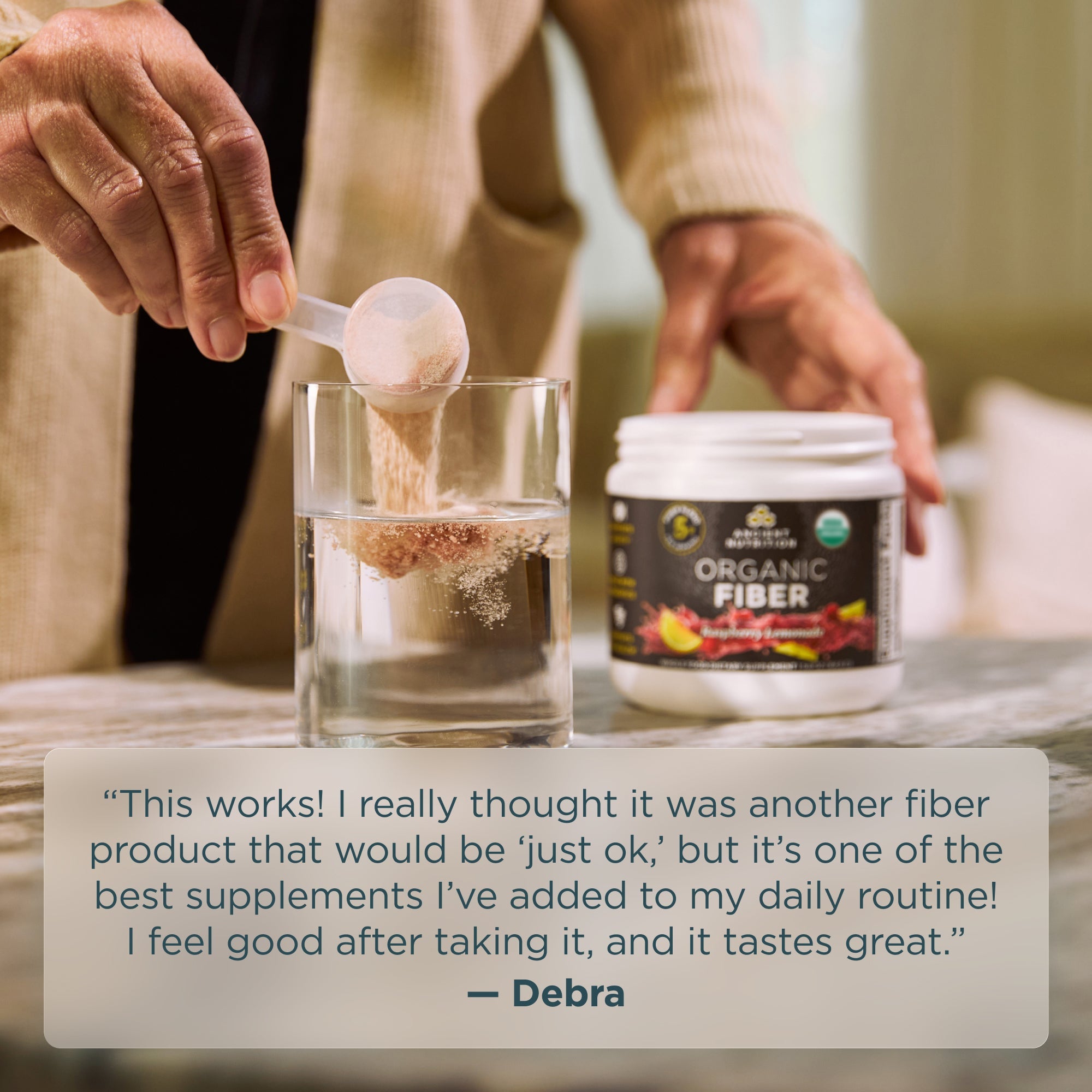 Person adding fiber powder to a glass with a container labeled 'Organic Fiber' on a table.