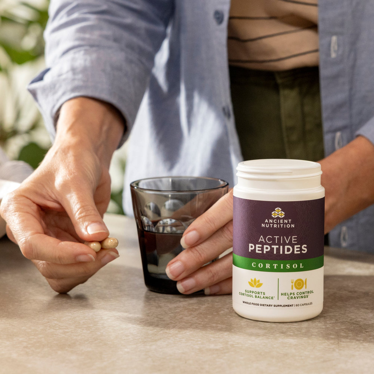 Person holding a supplement bottle labeled 'Active Peptides Cortisol' with a glass of liquid on a table.