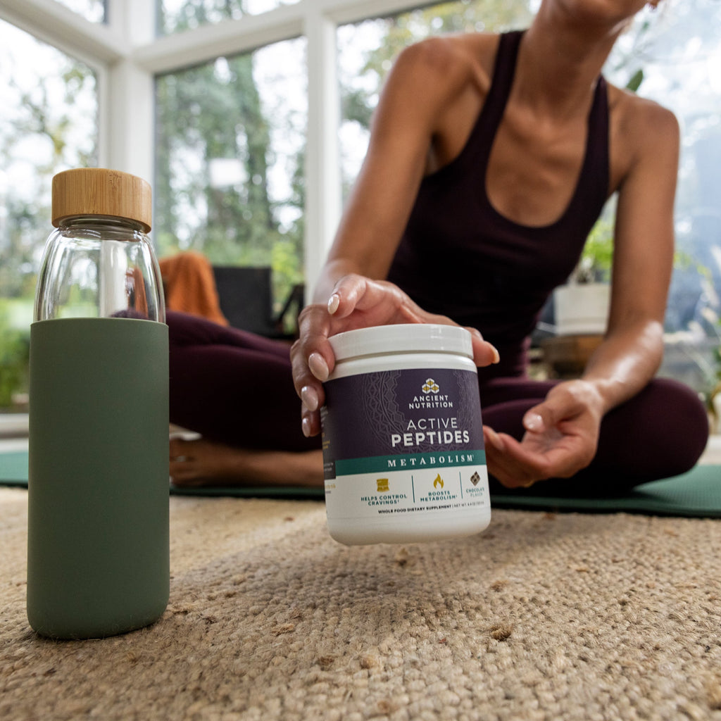 Person sitting on a yoga mat with a bottle and supplement container in the foreground
