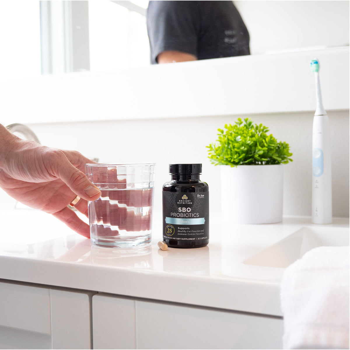 a man picking up a glass of water on a bathroom counter next to a bottle of  SBO Probiotics Men's capsules