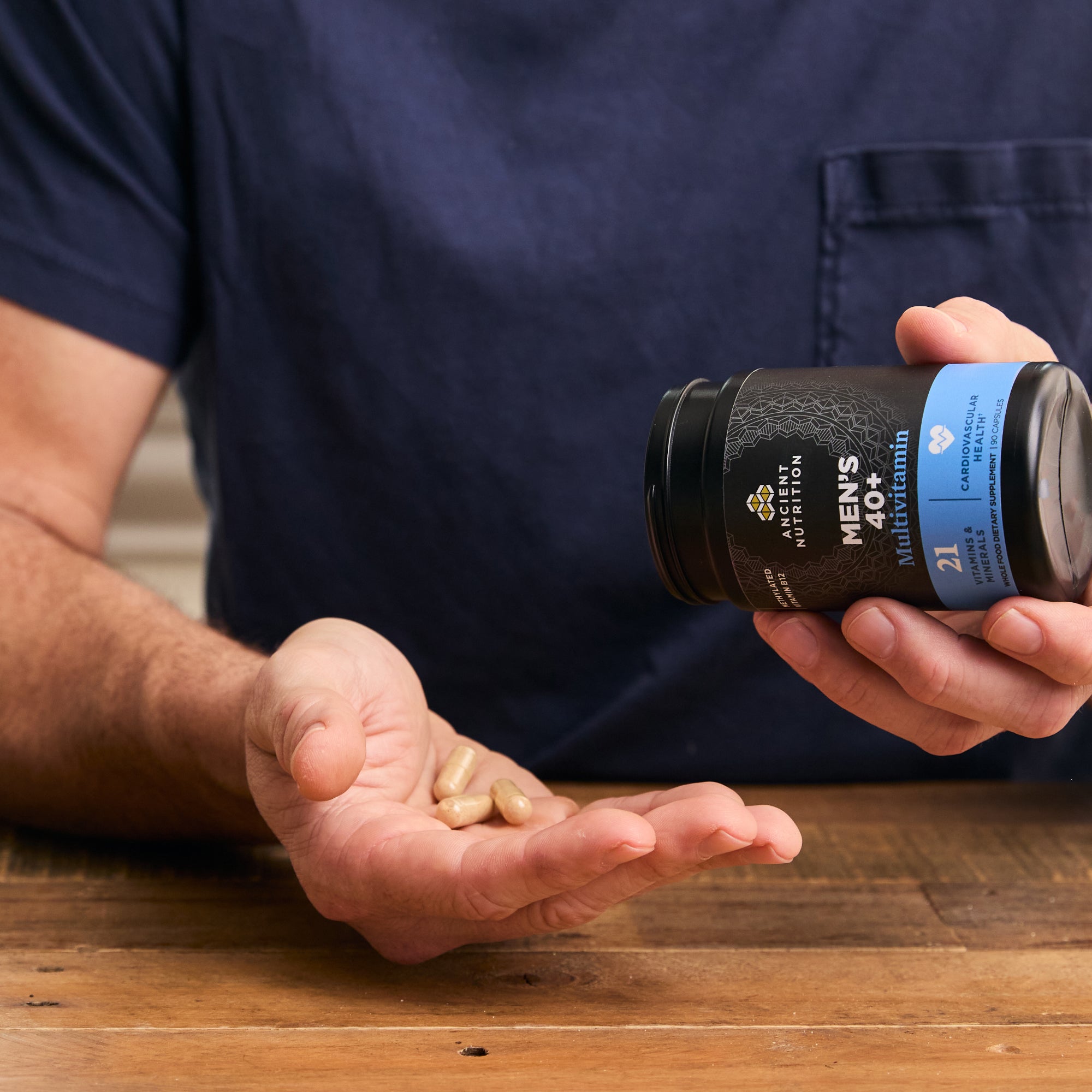 Person holding a bottle of Ancient Multivitamin Men's 40+ Capsules with pills in their hand on a wooden surface.