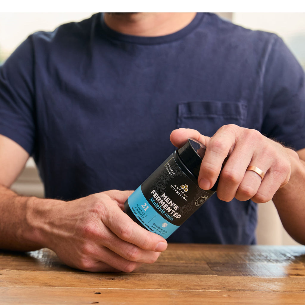Person holding a supplement container labeled 'Men's Fermented Multivitamin' on a wooden surface.
