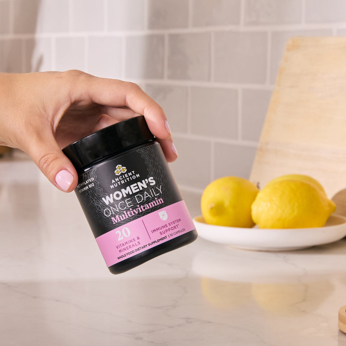 a person holding a bottle of Ancient Multivitamin Women's Once Daily Capsules (30 Capsules) with lemons in the background
